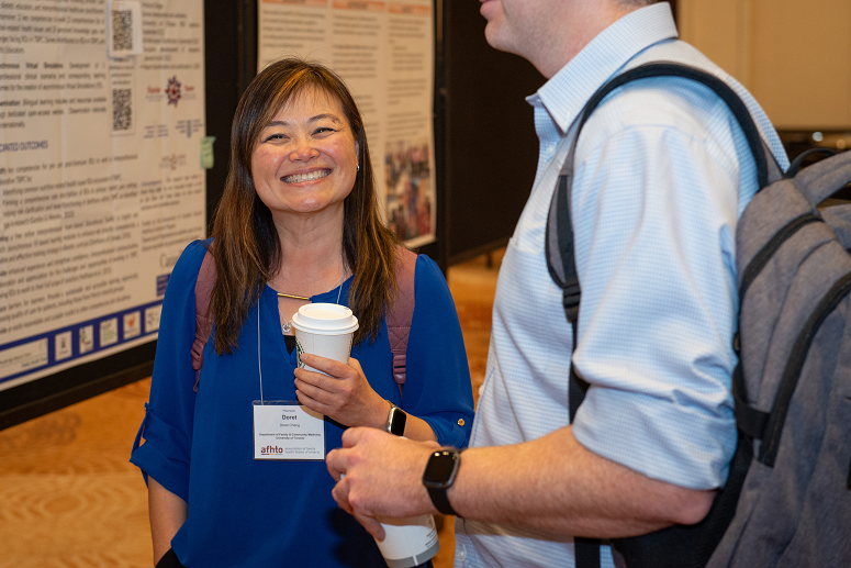 An Asian woman in blue top holding a coffee smiles at the camera as she stands in front of a poster with a colleague at the 2023 conference.