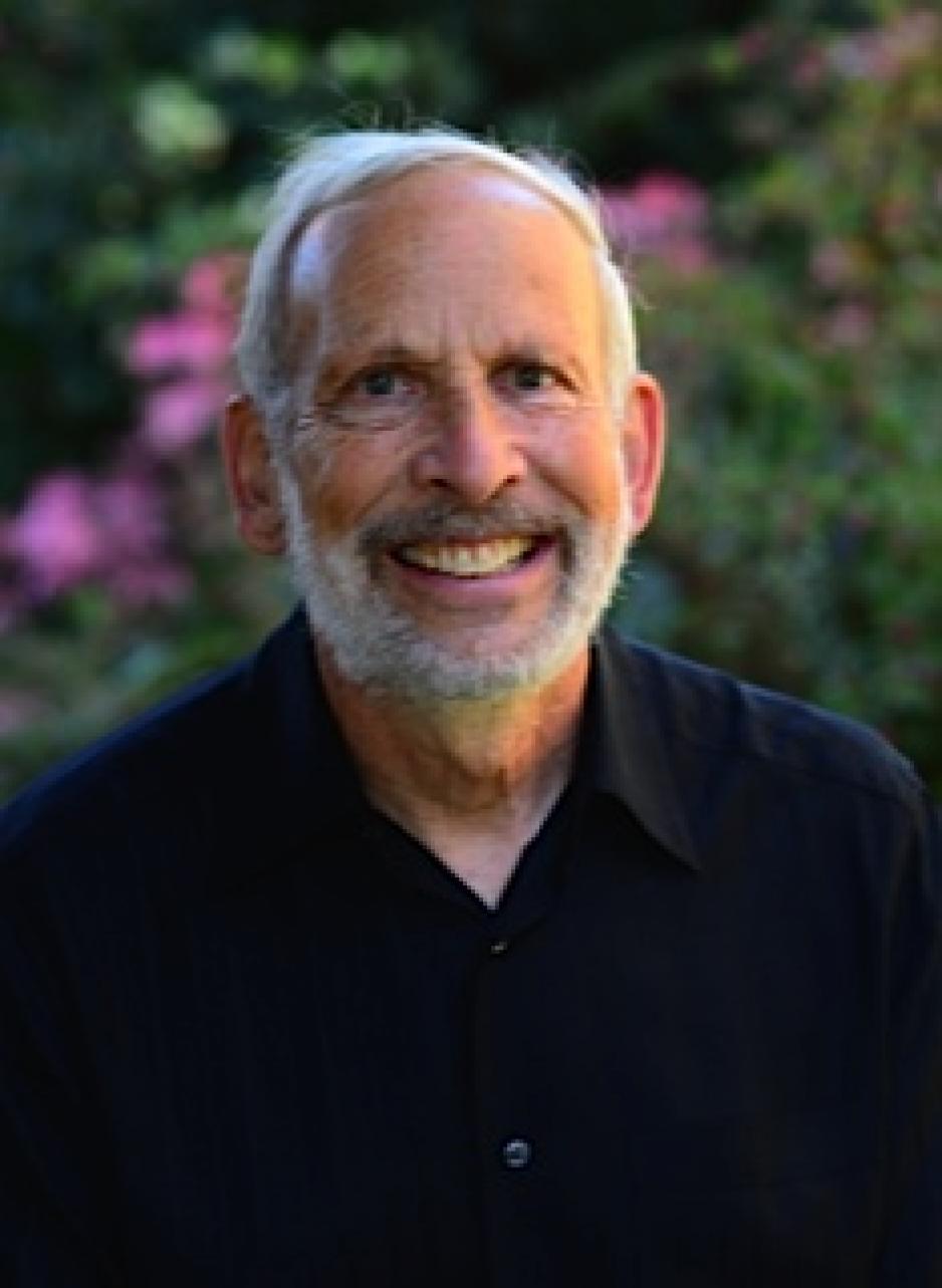 Dr. Thomas Bodenheimer has white hair and beard with grey mustache. He's wearing a black shirt and smiling. The background is blurred bushes with purple flowers.
